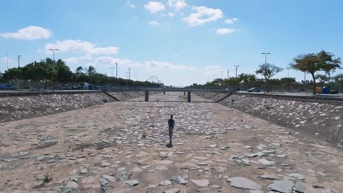 Young Man Walking Down the Drying River and Looking to Polluted City Carbon Dioxide on Background