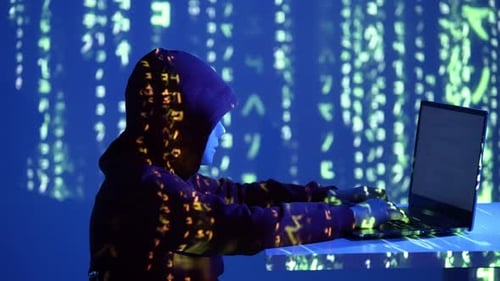 Un hacker anonyme portant un masque et un sweat à capuche utilise un ordinateur portable dans une pièce sombre avec projection de code Neon Matrix