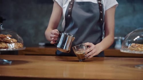 Front view of female worker that stands in cafe by the table and adds milk to coffee
