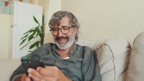 Close up, middle-aged man relaxing on his couch while using his smartphone to text