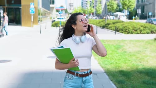 Caucasian Young Joyful Woman with Notebooks in Hand Walking in City and Speaking on Mobile Phone