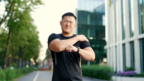Upset asian athletic man experiencing acute shoulder pain during morning run in urban city street.