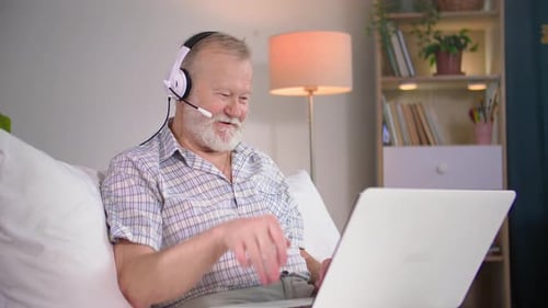Senior Man Chatting on Laptop with Headset at Home