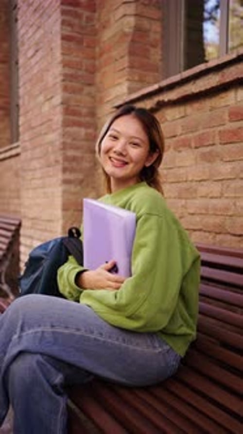 Portrait of Young Smiling Chinese Student Sitting on a Bench on Campus Looking at the Camera