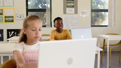 Diverse teenage classmates, boy responding to prompt, calling as girl typing on laptop in classroom