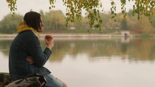 Young Handsome Man with Dreadlocks in Warm Clothes Drinking Coffee Near Lake in Golden Autumn