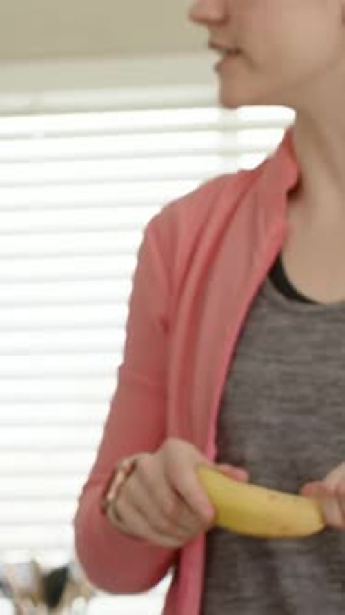 Vertical video: Turning left woman in coral zipup smartwatch peeling banana at kitchen, copy space