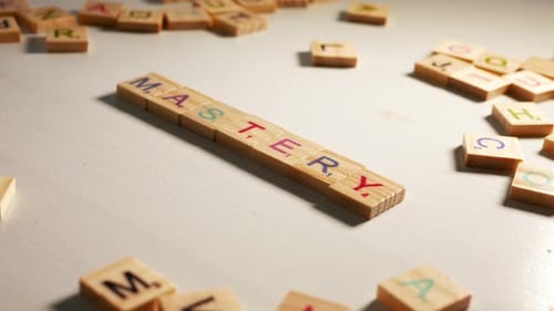 Someone writing Mastery with wooden tiles letters. Off white background. Dramatic light. Flat plane.