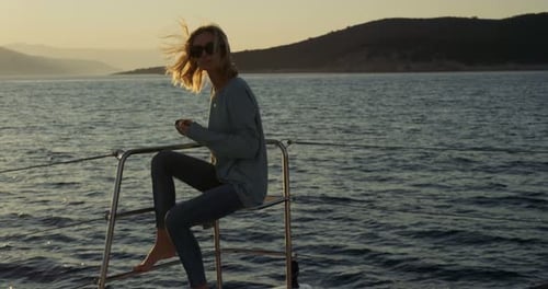 Beautiful Young Woman Loosens Her Blonde Hair While Sitting on a Yacht's Deck and Watching Sunset a