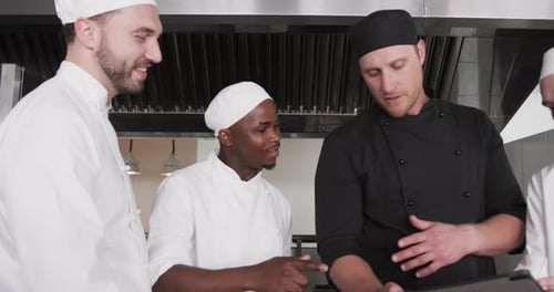 Diverse male chef instructing group of trainee male chefs using tablet in kitchen, slow motion