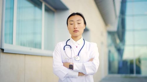 Close up portrait of Asian woman doctor looking at camera with arms crossed on background of modern