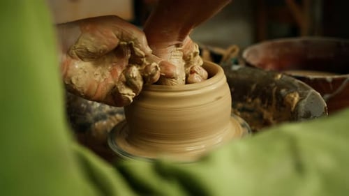 Close-up of potter's hands covered with clay making beautiful vase on throwing wheel in pottery