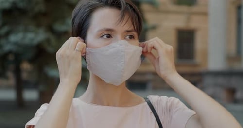 Portrait of Young Woman Putting on Medical Face Mask Then Turning to Camera Outdoors in City Street