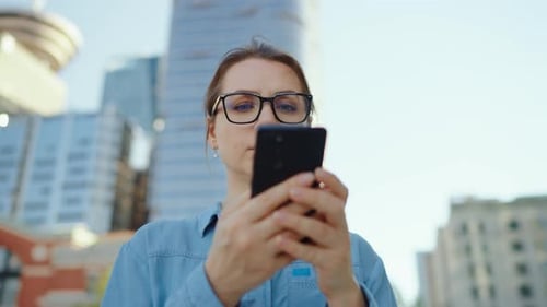 Woman in Glasses is Standing on the Street in the City and Using Smartphone