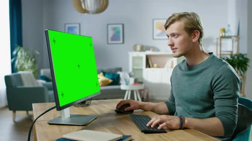Handsome Young Man Works on a Green Mock-up Screen Personal Computer while Sitting at His Desk in t