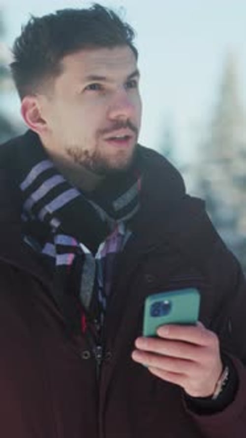 Person Wearing Winter Gear Focuses on Their Phone Surrounded By Snowy Conifers and Scenic Winter
