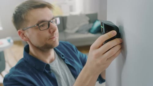 Young Man in Glasses Wearing a Blue Shirt is Hanging a Modern Movement Detector Unit on a White Wal