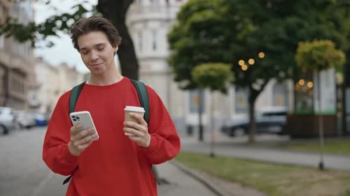 Handsome Caucasian Man with Brown Hair Holding Coffee Use Phone Smiling Walking on Street in City