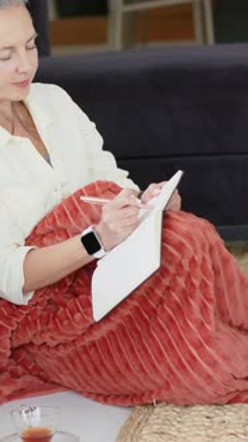 Vertical video: Camera panning revealing woman journaling in white pad on rug by sofa, rust throw