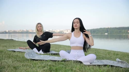 Two Young Women are Doing Yoga While Sitting on the Shore of the Lake