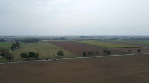 Aerial shot of rural road with driving cars surrounded by agricultural fields during cloudy day.