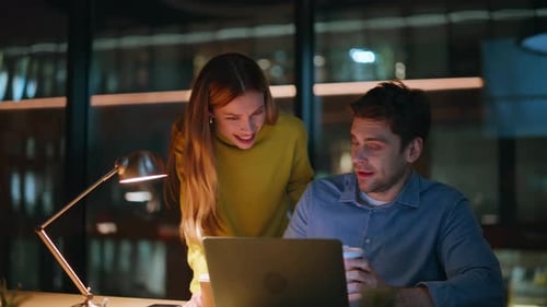 Lächelnde Kollegen diskutieren am späten Abend über einen Laptop im Startup-Look in Nahaufnahme
