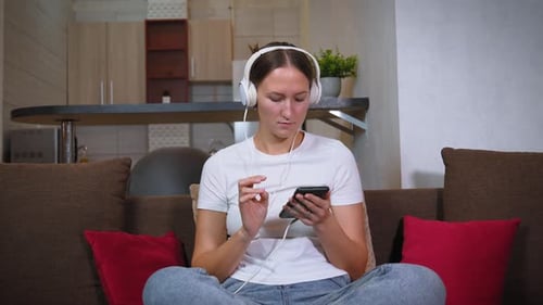 Junge Frau mit Kopfhörern surft auf ihrem Handy, während sie auf der Couch sitzt