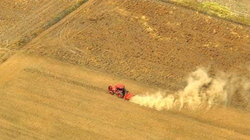 An aerial view shows a red tractor pulling a cultivator across a dry.