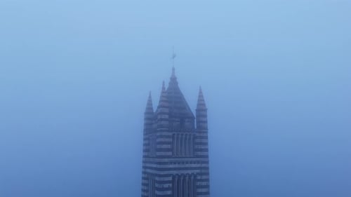 Siena Cathedral's Gothic tower emerges through dense fog in Tuscany, creating a mysterious scene