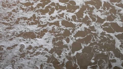 Aerial view of gentle waves creating frothy patterns as they wash onto a sandy beach. The intricate