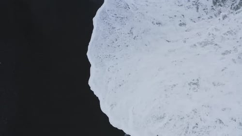 Aerial top view shot of foamy waves of ocean reaching black sand beach in Iceland