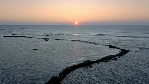Aerial flyover tranquil ocean water with outstanding rocks and golden falling sunset at horizon.