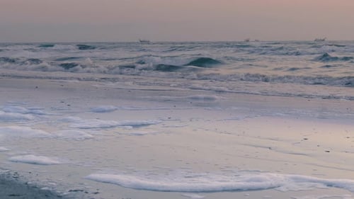 Waves gently wash over a peaceful sandy beach at sunset with foam scattered along the shore