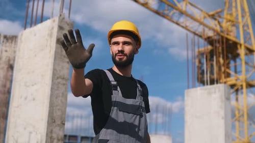 Showing stop gesture by hand. Young engineer in hard hat and uniform is on the construction site