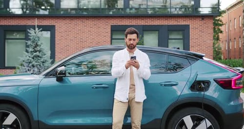 Man using smartphone near electric car charging outdoors