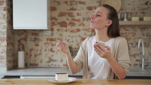 Woman Eats Cake While Using Smartphone in Kitchen