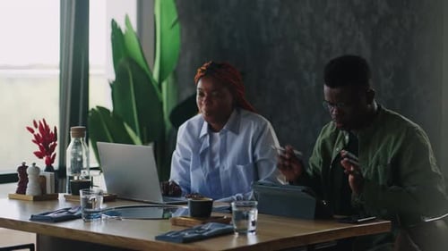 Two Black Employees Chat Over Cups of Coffee in a Local Cafe Reviewing a Business Plan on a Laptop