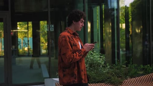 Smiling Man Walking on Street Near Modern Office Building Holding Phone Happy Browsing Internet News