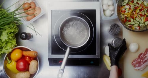 A woman in disposable black gloves adds salt to water boiling in a saucepan, close-up of her hands,