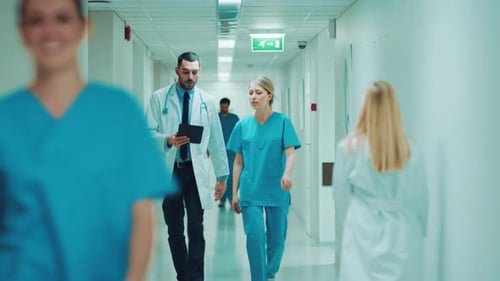 Female Surgeon and Female Doctor Walk Through Hospital Hallway, They Consult Digital Tablet Compute