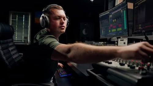 a focused male sound engineer wearing headphones sits at a mixing desk recording new track