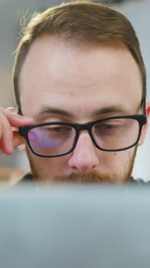 Likable Man Worker in Eyeglasses Sitting at the Table in Modern Office Room and Revisioning Details
