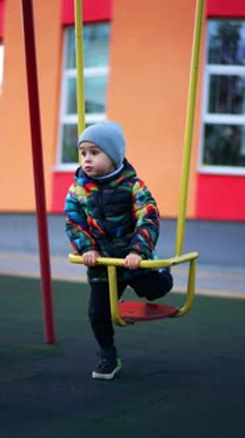 Toddler boy wearing warm clothes and cap sits on the swing.