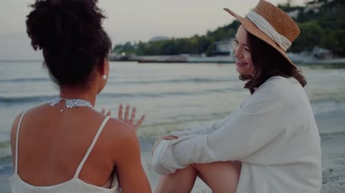 Two Females Sitting on Sandy Beach By Water at Warm Evening