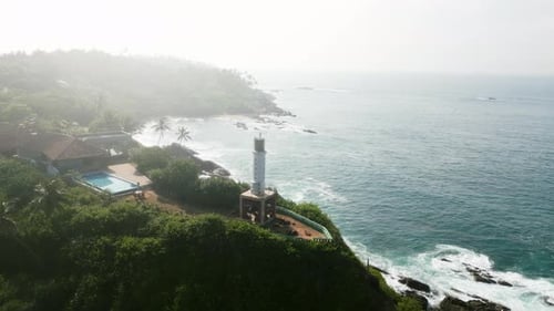 Aerial Video of Coastal Lighthouse on Cliff with Waves Crashing Below Green Trees Surround Historic