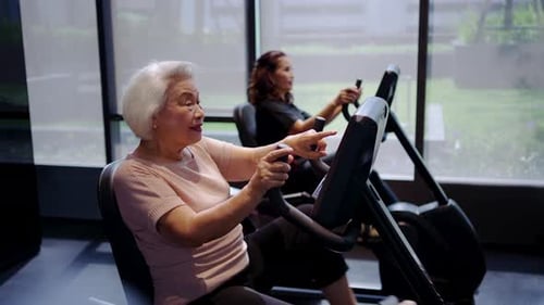 Two happy senior Asian women exercising on recumbent stationary bikes at gym, smiling and pointing