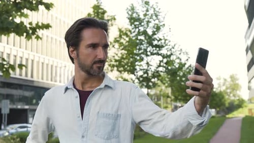 A Handsome Caucasian Man Takes Selfies with a Smartphone in an Urban Area