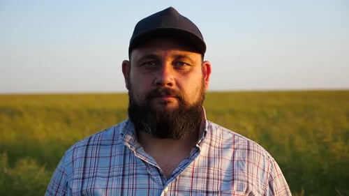 Un granjero serio con gorra mira a la cámara de pie ante un campo cultivado, retrato de un agrónomo adulto