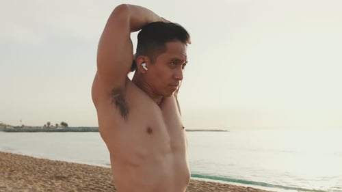 Young athletic man standing on the beach stretching his arm muscles before training
