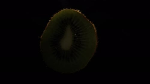 La mitad cortada del kiwi gira sobre un fondo negro. Primer plano. La luz se apaga lentamente.